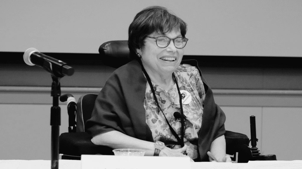 A photograph of Judith Heumann. She is sitting in her power chair behind table covered with a white table cloth. There is a microphone and a name placard in the foreground. She is wearing a colorful blouse and a maroon cardigan that matches the color of her glasses rims.